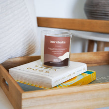 Load image into Gallery viewer, Candle labeled &#39;horchata&#39; on top of books in a wooden tray
